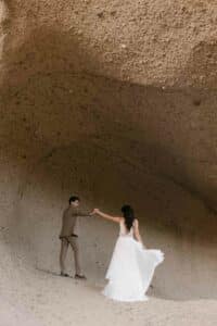 Im Herzen ihrer Hochzeit tanzt ein Paar anmutig in einer großen, in den Fels gehauenen Höhle. Der Bräutigam in einem braunen Anzug hält die Hand der Braut, deren fließendes weißes Kleid elegant wirbelt. Die natürliche Felsformation bietet eine intime Kulisse, mit glatten, geschwungenen Wänden, die ihre Liebesfeier umgeben.