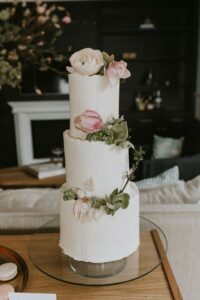 Eine wunderschöne dreistöckige weiße Torte mit glatter Glasur ist mit rosa und weißen Blumen geschmückt, perfekt für eine Hochzeit. Auf einem Glasständer in einem Wohnzimmer präsentiert, vervollständigt die Szene eine elegante Kulisse für die Hochzeitsreise eines jeden Paares.