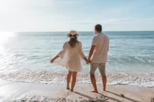 Ein Paar steht Hand in Hand an einem Sandstrand und träumt von seiner Hochzeit. Die Frau trägt ein weißes Kleid und einen Sonnenhut, während der Mann ein weißes Hemd und Shorts trägt. Sanfte Wellen umspülen ihre Füße unter dem klaren blauen Himmel, während sie über ihre gemeinsame Zukunft nachdenken.