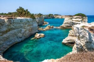 Klares türkisfarbenes Wasser umgibt schroffe weiße Klippen unter einem strahlend blauen Himmel – eine perfekte Kulisse für jede Hochzeitsplanung. Auf den Klippen wächst spärliches Grün, das sich im ruhigen Meer spiegelt. Der Blick fängt eine ruhige und malerische Küstenlandschaft ein.