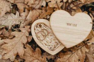 Eine herzförmige Holzbox mit der Gravur „wahre Liebe“ auf dem Deckel liegt offen auf einem Bett aus trockenem Herbstlaub. Perfekt für eine Hochzeit, in der Box befinden sich zwei Ringe auf einem Bett aus zerfetztem Papier, die den Beginn Ihrer schönen Reise in der Hochzeitsplanung symbolisieren.