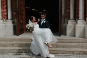 Ein Bräutigam im dunklen Anzug hält eine Braut im weißen Kleid auf den Stufen eines Gebäudes mit Holztüren und Backsteinmauern und verkörpert damit die Essenz ihrer Hochzeit. Die Braut, die Sandalen trägt und einen Blumenstrauß hält, lächelt ihn liebevoll an, während sie diesen schönen Moment gemeinsam genießen.