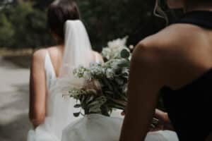 Eine Braut mit Schleier spaziert draußen umher, hält einen Strauß heller Blumen in der Hand und ist von der Schönheit der Natur umgeben. Neben ihr hält eine andere Person einen ähnlichen Strauß und fängt die Essenz einer Hochzeit in diesem grünen Paradies ein.
