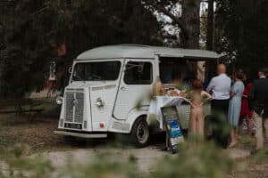 Ein weißer Vintage-Imbisswagen, perfekt für eine Hochzeit, parkt draußen, umgeben von Menschen an seinem Ausgabefenster. In dieser bezaubernden Szene, die sich ideal für Hochzeitsplanung eignet, schmücken Bäume und Bänke den Hintergrund, während die Gäste die Menütafel neben dem Wagen durchstöbern.