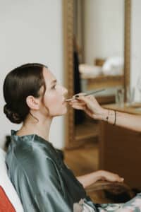 Eine Frau in einem Satinmantel sitzt da, während eine Visagistin gekonnt Lippenstift mit einem Pinsel aufträgt. Sie bereitet sich auf ihre Hochzeit vor. Sie ist drinnen und spiegelt sich in einem großen Spiegel mit verschwommenem Raumhintergrund. Ihr Haar ist ordentlich zu einem Knoten gestylt, was die Eleganz ihrer Hochzeitsplanung unterstreicht.