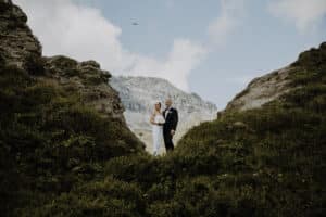 Braut und Bräutigam stehen zusammen auf einem üppigen, grünen Hügel inmitten schroffer Felsen. Die Szene ist perfekt für ihre Hochzeit, mit einer majestätischen Bergkette im Hintergrund unter einem teilweise bewölkten Himmel.