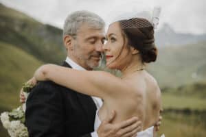 Ein Paar umarmt sich draußen in einer malerischen Landschaft, eingefangen in einem Moment perfekter Hochzeitsromantik. Die Frau in einem weißen Kleid und einem kleinen Hut mit Netz hält einen Blumenstrauß, während der Mann in einem dunklen Anzug sie sanft hält. Hügel und ein bewölkter Himmel bilden den Hintergrund für diese wunderschöne Hochzeitsszene.