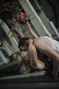 Zwei Menschen tanzen fröhlich, wobei einer den anderen in die Arme nimmt, was an einen perfekten Hochzeitsmoment erinnert. Sie halten einen Strauß roter Blumen, während im Hintergrund ein Gebäude mit großen Fenstern zu sehen ist.