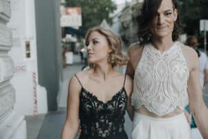 Zwei Personen schlendern in stilvollen Outfits eine Stadtstraße entlang, als ob sie gerade von einer Hochzeit zurückgekehrt wären. Die eine trägt ein schwarzes Spitzenkleid und die andere ein weißes Spitzentop. Beide haben hellbraunes Haar und wirken glücklich, während sie eng beieinander gehen, mit Bäumen und Gebäuden im Hintergrund.