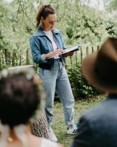 Eine Frau in Jeansjacke und Jeans steht draußen und liest in einem Buch, denkt vielleicht über ihre bevorstehende Hochzeit nach. Sie ist von Grün umgeben, im Hintergrund ist ein Holzzaun zu sehen. Im Vordergrund sind zwei Personen zu sehen, die etwas unscharf sind.