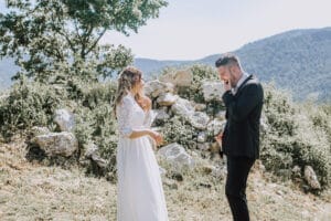 Eine Braut im weißen Kleid und ein Bräutigam im schwarzen Anzug stehen draußen und lächeln sich in der atemberaubenden Berglandschaft an, perfekt für ihre Hochzeit. Umgeben von Grün und Felsen, unter dem klaren, sonnigen Himmel, ist es eine ideale Umgebung, damit ihre Liebe gedeihen kann.