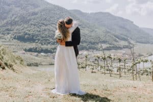 Braut und Bräutigam umarmen sich auf einem grasbewachsenen Hügel mit Blick auf ein malerisches Tal und halten so einen perfekten Hochzeitsmoment fest. Die Braut trägt ein weißes Kleid und hält ihren Blumenstrauß, während der Bräutigam in einem dunklen Anzug dasteht. Üppige grüne Hügel und ein Fluss erstrecken sich unter dem klaren Himmel – ein wahrer Beweis für sorgfältige Hochzeitsplanung.