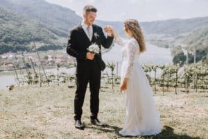 Ein Paar in Hochzeitskleidung steht auf einem grasbewachsenen Hügel und genießt die Freude seiner Hochzeit. Der Bräutigam im schwarzen Anzug hält die Hand der Braut, die ein weißes Kleid mit Spitzenärmeln trägt. Gemeinsam genießen sie diesen freudigen Moment vor der Kulisse einer malerischen Fluss- und Berglandschaft.