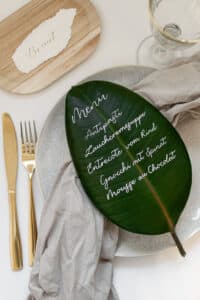 Ein kreatives Menü, perfekt für eine Hochzeit, ist mit weißer Tinte auf ein großes grünes Blatt geschrieben. Es umfasst Antipasti, Suppe, Rinderentrecôte, Spinat-Gnocchi und Schokoladenmousse. Es ist elegant auf einem weißen Teller mit einer beigen Serviette platziert, neben goldenem Besteck und einem Weinglas.