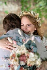 Eine Frau mit roten Haaren und einem Blumenkranz umarmt freudig eine Person in einem hellgrauen Anzug. Sie hält einen Blumenstrauß in zarten Rosa- und Weißtönen, der den Geist ihrer Hochzeit verkörpert. Grün und ein unscharfer natürlicher Hintergrund verleihen diesem Hochzeitsmoment eine heitere Atmosphäre.