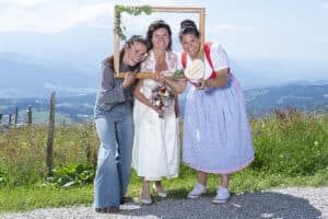 Drei Frauen posieren draußen vor einer Bergkulisse während einer wunderschönen Hochzeit. Eine hält einen Holzrahmen um die Braut, während eine andere ein herzförmiges Schild hält. Die Braut bereitet sich auf die Hochzeit vor, hält einen Blumenstrauß und trägt ein weißes Kleid. Sie alle lächeln unter dem klaren blauen Himmel.