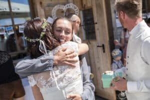Bei einer Hochzeit im Freien umarmt eine Braut in einem weißen Spitzenkleid und mit Blumenkopfschmuck eine Frau in einem grauen Hemd. Ein Mann in einer weißen Weste hält eine Geschenkbox und ein Glas. Gäste und Dekorationen schaffen die perfekte Kulisse für dieses besondere Fest der Liebe und sorgfältigen Hochzeitsplanung.