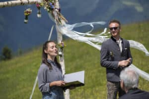 Eine Frau und ein Mann stehen draußen vor einem dekorierten Bogen mit Bergen im Hintergrund und feiern ihre Hochzeit. Die Frau hält einen Laptop, während der Mann mit der Sonnenbrille lächelt. Einige Gäste sitzen davor, teilweise sichtbar, was den Charme dieses unvergesslichen Moments noch verstärkt.