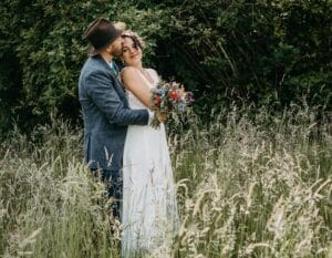 Ein Paar umarmt sich auf einem Feld mit hohem Gras. Die Frau, in einem weißen Kleid, das perfekt zu ihrer Hochzeit passt, hält einen bunten Blumenstrauß. Der Mann, mit Hut und Jacke, küsst sie auf die Stirn. Üppiges grünes Laub bildet den Hintergrund und schafft eine ruhige, natürliche Umgebung, ideal zum Heiraten.