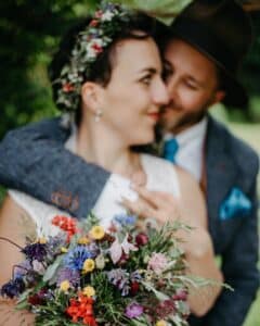 Ein Paar umarmt sich im Freien und verkörpert eine verträumte Hochzeitsszene. Der Mann in Hut und Anzug hält sanft die mit einer Blumenkrone geschmückte Frau, während sie einen leuchtend bunten Blumenstrauß umklammert. Üppiges Grün dient als perfekte Kulisse für diesen bezaubernden Moment.