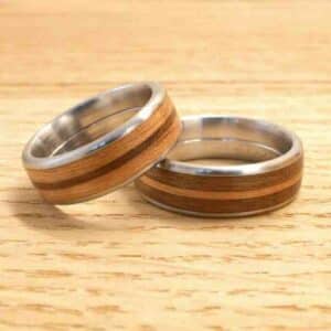 Zwei silberne Ringe mit Holzeinlagen liegen auf einer Holzoberfläche und verkörpern ein rustikales und doch modernes Design. Die glatten Metallkanten rahmen das zentrale Holzband ein, perfekte Symbole für eine einzigartige Hochzeit oder für diejenigen, die sich intensiv mit der Hochzeitsplanung beschäftigen.