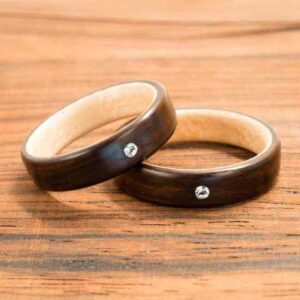 Zwei Holzringe mit kleinen eingelegten Edelsteinen liegen auf einer Holzoberfläche, perfekt für Ihre Hochzeit. Die Ringe haben eine glatte, dunkle Oberfläche und eine helle Innenseite, ideal für Paare mitten in ihrer Hochzeitsplanung.