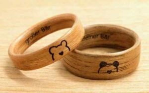 Zwei Holzringe auf heller Oberfläche, in die jeweils ein Bärengesicht und ein kleines Herz eingraviert sind. Perfekt für jede Hochzeit: Auf einem Ring steht „großer Bär“, auf dem anderen „kleiner Bär“, was Ihrer Hochzeitsplanung einen sentimentalen Charme verleiht.