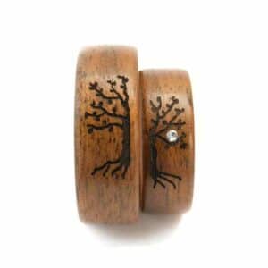Zwei Holzringe, perfekt für die Hochzeitsplanung, sind mit Baummotiven graviert. Ein Ring ist mit einem kleinen Edelstein verziert, der in den Baum eingelassen ist. Sie liegen elegant nebeneinander auf einem weißen Hintergrund und fangen die Essenz des Heiratens ein.