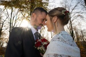 Ein Paar im Hochzeitskleid steht dicht beieinander und lächelt sich herzlich an. Der Bräutigam hält einen Strauß roter und weißer Blumen in der Hand. Sonnenlicht fällt durch die Bäume und schafft eine strahlende Hochzeitsplanungsatmosphäre.
