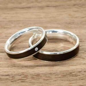 Zwei silberne Ringe mit schwarzen Einlagen und einem kleinen Diamanten darin, nebeneinander auf einer Holzoberfläche platziert, fangen die Essenz der Hochzeitsplanung ein.