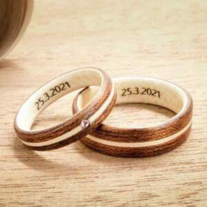 Zwei Holzringe mit eingraviertem Datum „25.3.2021“ werden auf einer Holzoberfläche präsentiert, perfekt für alle, die gerade mitten in der Hochzeitsplanung stecken. Ein Ring ist mit einem kleinen Edelstein versehen, der dem schlichten, natürlichen Design eine elegante Note verleiht, ideal für alle, die eine Hochzeit mit Stil und Anmut planen.