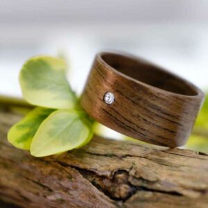Ein Holzring mit einem kleinen Edelstein ist auf einem Stück Treibholz platziert. Grüne Blätter im Hintergrund sorgen für eine natürliche und erdige Kulisse, perfekt als Inspiration für diejenigen, die gerade mitten in der Hochzeitsplanung stecken.