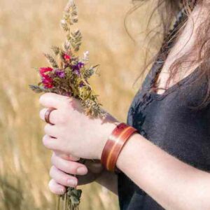 Eine Person hält einen Strauß Wildblumen, geschmückt mit einem Holzarmband und einem Ring, und denkt vielleicht über ihre Hochzeitsplanung nach. Mit langen, welligen braunen Haaren und einem schwarzen Oberteil steht sie an diesem sonnigen Tag vor der goldenen Feldkulisse.