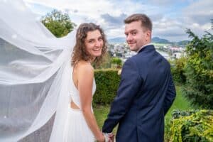 Ein fröhliches Paar im Hochzeitskleid steht draußen und hält Händchen, was die Aufregung ihrer Hochzeit ausdrückt. Der Schleier der Braut weht im Wind, während beide in die Kamera lächeln. Ein malerischer Hintergrund mit Bäumen und einer Stadtlandschaft vor Hügeln entfaltet sich unter einem teilweise bewölkten Himmel und fängt ihren perfekten Tag ein.