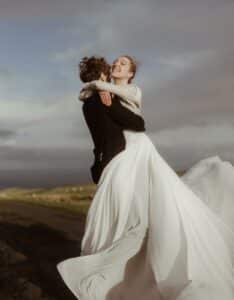 Ein glückliches Paar umarmt sich im Freien, wobei die Frau im fließenden weißen Kleid von ihrem Partner im dunklen Anzug hochgehoben wird. Der Himmel ist bedeckt und schafft eine dramatische Kulisse – ein bezaubernder Moment auf ihrer Hochzeitsreise.