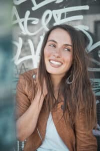 Eine Frau mit langen braunen Haaren lächelt, während sie in einer braunen Lederjacke und einem weißen Oberteil dasteht. Ihre Begeisterung für die Hochzeitsplanung ist vor der mit farbenfrohen Graffiti bedeckten Wand deutlich zu erkennen.