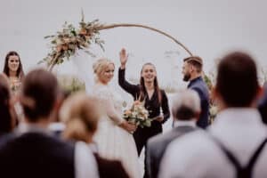 Braut und Bräutigam stehen bei ihrer Hochzeit im Freien zusammen. Der lächelnde Geistliche hebt beim Sprechen die Hand. Die Braut hält einen Blumenstrauß und im Vordergrund sind Gäste zu sehen. Ein mit Blumen geschmückter Bogen ziert den Hintergrund und zeigt exquisite Hochzeitsplanung.