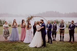 Braut und Bräutigam küssen sich unter einem runden Blumenbogen in einer nebligen Umgebung im Freien und verkörpern damit den Geist einer Hochzeit. Umgeben von Brautjungfern in farbenfrohen Kleidern und Trauzeugen in Anzügen und traditioneller Kleidung stehen sie auf einer Rasenfläche in der Nähe des Sees und spiegeln eine sorgfältige Hochzeitsplanung wider.