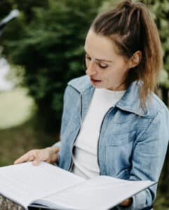 Eine Frau mit Pferdeschwanz, die wahrscheinlich über ihre eigene Hochzeitsplanung nachdenkt, liest im Freien ein Buch. Sie trägt eine Jeansjacke über einem weißen Hemd, umgeben von üppigem Grün, das an eine ruhige Garten- oder Parklandschaft erinnert.