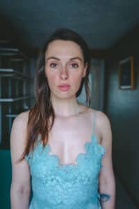 Eine Frau mit langen braunen Haaren und einem blauen Spitzentop steht drinnen und verkörpert die Essenz von Eleganz. Sie blickt aufmerksam in die Kamera, als würde sie über ihre bevorstehende Hochzeit nachdenken. Der schwach beleuchtete Raum hinter ihr ist mit Regalen und einem gerahmten Bild an der Wand ausgestattet, was eine intime Atmosphäre schafft.