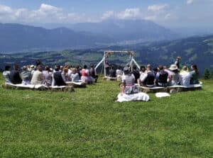 Eine Gruppe von Menschen sitzt auf Heuballen auf einem offenen Feld und nimmt an einer Hochzeitszeremonie teil. Vorne ist ein dekorativer Bogen aufgestellt, der einen malerischen Blick auf Berge und ein Tal unter einem teilweise bewölkten Himmel bietet.
