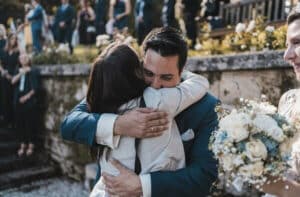Ein Mann im blauen Anzug umarmt herzlich eine Frau in einem hellen Outfit und feiert ihre Hochzeit. Eine weitere Person mit einem Strauß weißer Rosen steht daneben. Im Hintergrund, teilweise hinter einer Steinmauer sichtbar, sind Gäste in die Freude der Hochzeitsfeierlichkeiten versunken.