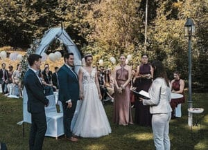 Eine wunderschöne Hochzeit im Freien findet statt, während das Paar unter einem Bogen steht. Die Braut glänzt in einem weißen Kleid, während der Bräutigam in einem dunklen Anzug schick aussieht. Brautjungfern in rosa Kleidern stehen in der Nähe und die Gäste sitzen rundherum. Bäume und Luftballons verleihen dieser perfekt geplanten Hochzeitsplanung einen besonderen Charme.