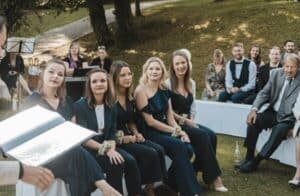 Eine Gruppe von Menschen sitzt im Freien bei einer Hochzeitszeremonie. Im Vordergrund sitzen fünf Frauen in Abendgarderobe auf einer Bank und halten kleine Blumensträuße in den Händen. In der Nähe sitzen weitere Gäste auf weißen Bänken zwischen Bäumen und Gras, was die sorgfältige Hochzeitsplanung unterstreicht.