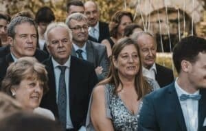 Eine Gruppe von Menschen in formeller Kleidung sitzt im Freien bei einer Hochzeitsfeier. Die Gäste wirken verlobt, manche lächeln und genießen den Moment. Im Hintergrund funkeln Lichterketten, die zur festlichen Atmosphäre beitragen und eine wunderbare Hochzeitsplanung zeigen.