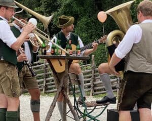 Eine Gruppe von Männern in traditioneller bayerischer Kleidung spielt während einer Hochzeit im Freien Instrumente, darunter Tuba, Gitarre, Trompete und Posaune. Sie versammeln sich um Notenständer mit einem Holzzaun und Grün im Hintergrund. Einige Bierflaschen sind zu sehen.
