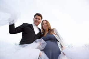 Ein Mann in schwarzem Anzug und weißem Hemd steht neben einer Frau in blauem Kleid und weißem Schal und fängt die Essenz der Hochzeitsplanung ein. Sie halten weißes Tuch und blicken in die Kamera, vor einem hellen, bewölkten Himmel – ein perfekter Moment zum Heiraten.
