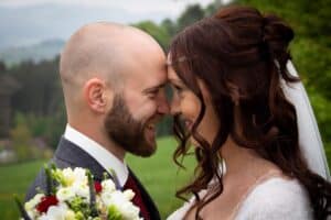 Braut und Bräutigam sind in der Freude ihrer Hochzeit versunken und lächeln sich an, während sie ihre Stirnen in einer ruhigen Umgebung im Freien berühren. Die Braut umklammert einen farbenfrohen Blumenstrauß, während sie in eleganter Hochzeitskleidung stehen, umgeben von üppigem Grün und Bäumen.