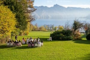 Eine Gruppe von Menschen sitzt in Reihen im Freien auf einer Wiese und blickt auf einen Redner. Ein großes weißes „JA“ auf dem Rasen deutet auf eine elegante Hochzeit hin. Im Hintergrund tragen ein ruhiger See und Berge unter einem klaren Himmel zum Charme bei. Bäume mit Herbstlaub vervollständigen diese malerische Kulisse.