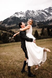 Ein fröhliches Paar im Hochzeitskleid feiert seine Hochzeit auf einer Wiese. Der Bräutigam hebt die Braut hoch, die einen Blumenstrauß hält. Beide lächeln vor einer Kulisse aus schneebedeckten Bergen und bewölktem Himmel.